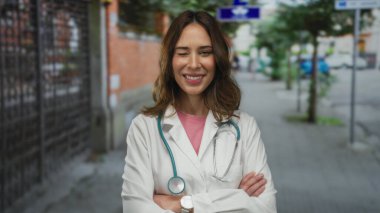 Steteskoplu İspanyol kadın doktor şehir sokaklarında kollarını kavuşturup açık hava ortamında profesyonelliği tasvir ediyor..