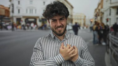 Meşgul bir sokakta göğsünü açık havada tutan sakallı genç bir adam, rahatsızlığını dile getiriyor, çizgili gömlek giyiyor, kentsel bir ortamda sağlık sorunları yaşıyor..