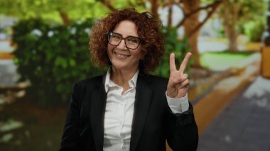 Parlak park ortamında gülümseyen bir kadın neşeli bir ifade sergiliyor. Açık havada olumlu bir jest yapıyor. Yeşillik ağaçlar ve patikalarla arka planı vurguluyor.