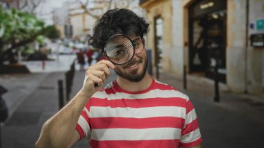 Gözlüklü, sakallı, elinde büyüteç olan kırmızı çizgili gömlek giyen, şehir ortamında gezen Latin kökenli genç bir adam..