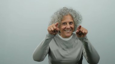 Gri saçlı bir kadın iki eliyle buraya gelir izole edilmiş beyaz bir arka plan duvarına gülümser, dostça bir davetkar tavır sergiler..