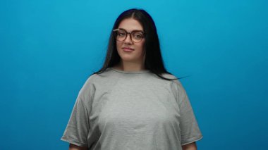 Gözlüklü kadın, sağlam mavi bir arkaplan karşısında uzun saçlı, kıvrımlı ve günlük gri gömlekli bir kadın pozu veriyor..