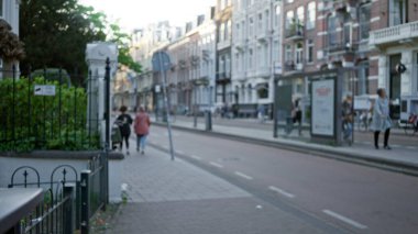 Amsterdam caddesinde yürüyen, dağılmış binalar ve ağaçlar üzerinde yürüyen, Hollanda 'daki kentsel yaşamı bokeh etkisi altında yakalayan insanların bulanık görüntüsü..