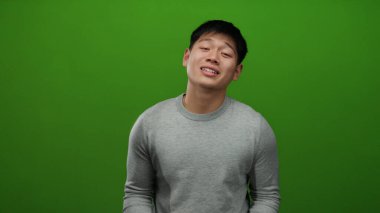 Parlak yeşil arkaplan önünde güven içinde el kol hareketi yapan, günlük gri süveter giyen, duygularını basit bir stüdyo arkaplanına karşı elleriyle ifade eden genç adam.