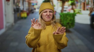 İğrendiğini ifade eden sarı kazak ve bereyle şehir caddesinde duran bulanık geçmişli bir kadın.