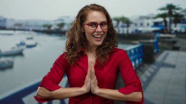 Sahilde gezinen kırmızı tişörtlü bir kadın. Arka planda bulanık botlar ve binalar, güneşli bir plaj gününde neşe ve rahatlama somutlaştırıyor..