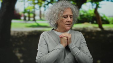 Gri saçlı, güneşli bir park ortamında dalgın dalgın duran yaşlı bir kadın..