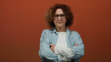 Orta yaşlı esmer, esmer ve gözlüklü, esmer, esmer bir kadın kollarını düz kahverengi bir duvara yaslamış, sıradan bir kot gömlek giyiyor..