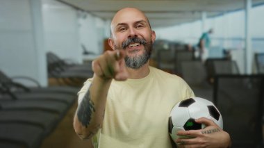 Futbol topu tutan ve parlak gökyüzünün altında bir yolcu gemisinde duran sakallı neşeli kel adam.