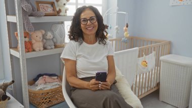 Bebek beşiği ve pelüş oyuncaklarla rahat yatak odasında cep telefonu kullanan olgun İspanyol kadın, sıcak bir iç mekan ortamı yaratıyor..