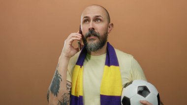 Telefondan konuşan sakallı bir adam futbol topunu tutuyor ve kahverengi arka planda eşarp takıyor.