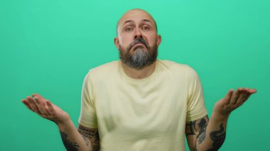 Sarı gömlekli, sakallı ve dövmeli bir adam yeşil arka plana karşı omuz silkiyor..