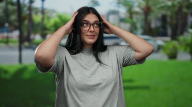 Parkta gözlüklü bir kadın poz veriyor, büyük beden bir figür sergiliyor, yeşillik ve şehir zemininin ortasında duruyor, duygularını ifade ediyor, uzun siyah saçlar ve günlük gri bluzuyla..