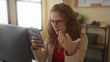 Ofiste akıllı telefon kullanan kafası karışık bir kadın, kırmızı gözlük takıyor, modern bir iç mekanı olan bir masada oturuyor, iş iletişimi veya teknoloji kullanımını öneriyor..