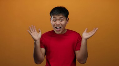 İzole edilmiş turuncu duvarın üzerinde kırmızı tişört giyen genç adam çeşitli duyguları ifade ediyor. Yüzünde eller ve hareketler var..