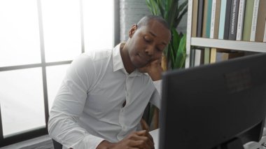 Ofisteki genç adam, beyaz gömlekli, düşünceli bir şekilde bilgisayara bakıyor. Kafasını kitaplıklara yaslıyor ve odada pencere ışığı yanıyor.