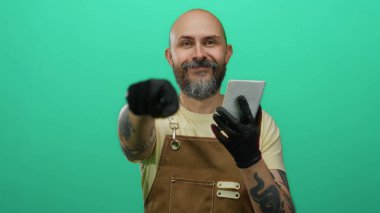 Sakallı ve dövmeli bir adam yeşil arka plana karşı defter tutarken önlük ve eldiven takıyor..
