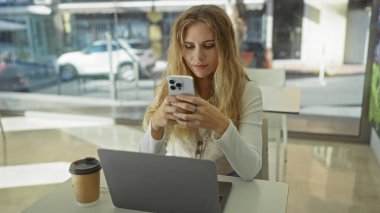 İş yerinde bilgisayarlı ve kahveli akıllı telefon kullanan bir kadın modern ofis ortamında sokak manzaralı, mobil iletişime odaklanmış sarışın bir yetişkin..