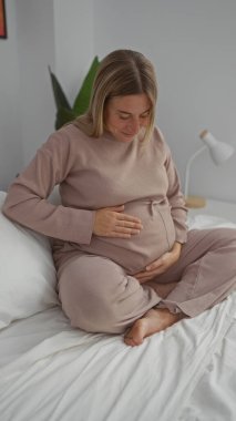 Sarı saçlı hamile bir kadın, rahat bir yatak odasında, karnına hafifçe dokunarak, hamileliğin güzelliğini kucaklayarak, doğal ışıkla, huzurlu bir atmosfer yaratıyor..