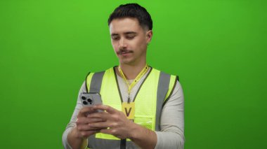 Yansıtıcı yelek giyen genç İspanyol adam, gönüllü ortamında, odaklanmış ve nişanlı olarak izole edilmiş yeşil arka plana karşı akıllı telefon tutuyor..
