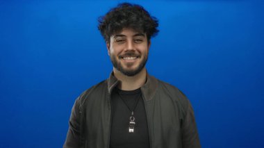 Sakallı, parlak mavi arka planda dikilen, rahat ve kendinden emin bir tavır takınan, siyah ceketli, Latin asıllı genç bir adam..