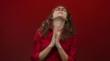 Kırmızı arka plan karşısında gözlük takan kadın, izole edilmiş alanda duygu ve inanç gösteren umut dolu bir ifadeyle dua ediyor..