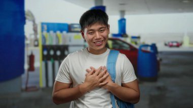 Bir benzin istasyonunda göğsü açık gülümseyen genç bir adam, beyaz gömlek ve mavi bir sırt çantası giyiyor, parlak kırmızı arabanın ve benzin pompalarının önünde duruyor..