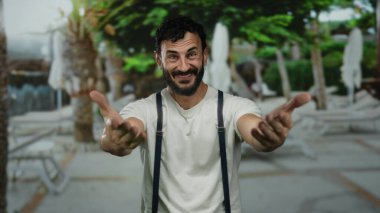 Sakallı İspanyol bir adam güneşli bir sokakta arka planda palmiye ağaçları olan beyaz bir gömlek ve pantolon askısı takarak coşkuyla el kol hareketi yapıyor..