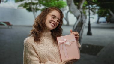 Dışarıda elinde pembe kurdeleye sarılı bir hediye kutusuyla gülümseyen bir kadın, şehir ortamında mutluluğu gösteriyor..