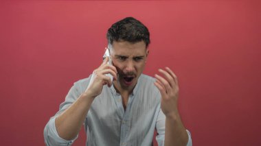 Genç İspanyol asıllı bir adam, izole edilmiş kırmızı arka planda akıllı telefondan konuşuyor yoğun jestlerle hayal kırıklığını ifade ediyor, hararetli bir sohbeti işaret eden sıradan bir gömlek giyiyor..