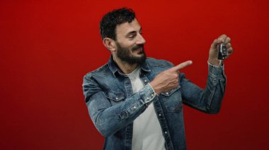 Kot ceketli İspanyol adam heyecan verici bir şekilde elinde kırmızı arka planlı araba anahtarı tutuyor. Promosyon amaçlı dinamik ve canlı bir atmosfer yaratıyor..