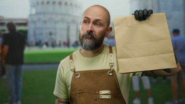 Kahverengi önlüklü kel adam güneşli bir günde arka planda sidik kulesi olan bir kesekağıdı tutuyor..