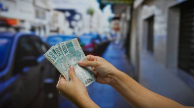 Bir şehir sokağında Brezilya banknotları tutan bir adam, açık bir şehir ortamında iş alım satımı teması taşıyor..