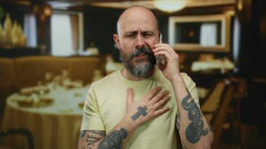 Restoranda boğazında ve telefonunda dövmeler olan sakallı bir adam rahatsız ve endişeli görünüyor..