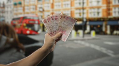 Elinde Türk lirası tutan bir adam, arka planda şehir sokağı ve kırmızı otobüsle açık havada, kalabalık bir metropol ortamında şehir hayatı ve finans temalarını gözler önüne seriyor..