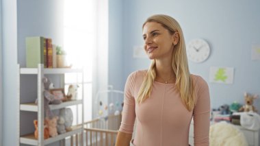 Oyuncak ve beşiğiyle rahat bir bebek odasında duran, doğal ışıkla gülümseyen, dingin ve hoş bir atmosfer yaratan bir kadın..