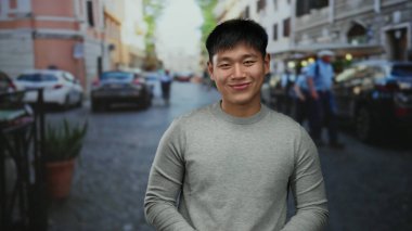 Kalabalık bir şehir caddesinde gülümseyen genç adam, neşeli ve huzurlu bir ruh halini yansıtıyor. Arabalar ve binalar arasında, gri bir süveter giyiyor..