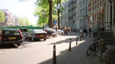 Hollanda, Amsterdam 'da, güneşli bir kanal boyunca bulanık yayalar ve arabalarla, ikonik Hollanda mimarisi ve canlı bir şehir atmosferi gösteren sokak manzarası..
