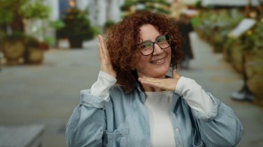 İspanyol kadın hareketli bir şehir caddesinde neşeli jestler yapıyor, canlı bitkiler ve hafif ışıklandırmayla çerçevelenmiş, canlı bir açık şehir anını yakalıyor..