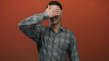 Kalıplı gömlekli genç İspanyol adam, izole edilmiş kırmızı arka plana karşı elleriyle gözlerini kapatıyor..