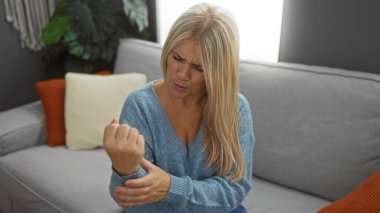 İçerideki kadın rahat bir oturma odasında kol ağrısı çekiyor, şık bir dekorla çevrili, endişeli bir ifade sergiliyor, yetişkin kadın güzelliğini ve aile rahatlığını somutlaştırıyor..