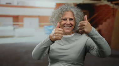 Kapalı bir inşaat sahasında baş parmaklarıyla gülümseyen bir kadın, kendine güveni ve pozitifliği olan kıvırcık gri saçlı bir kadın..