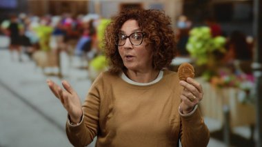 Şehir caddesinde elinde kurabiye tutan orta yaşlı İspanyol bir kadın. İnsanlarla canlı bir arka plan ve bulanık aktiviteler..