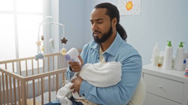 Sakallı bir adam bebeği yatak odasında tutuyor ve sıcaklığı dijital termometreyle ölçüyor. Sıcak, şefkatli bir ortam yaratıyor..