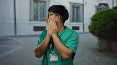 Tıbbi önlüklü genç adam şehir caddesinde dikiliyor elleri ağzında ürkmüş görünüyor.