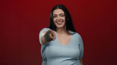 Gülümseyen ve parlak kırmızı arka plana işaret eden bir kadın, özgüveni ve pozitifliği somutlaştıran, bir stüdyoda geçen, açık mavi bir bluzla artı boy temsil eden bir kadın..