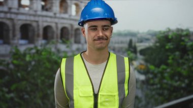 Yansıtıcı yelek giyen genç adam Roma Kolezyumu 'nun yakınında duruyor. Kendine güvenen bir ifadeyle tarihin ve modern yapının bir karışımını yakalıyor..