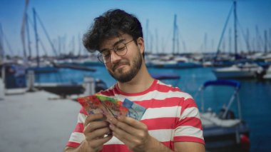 Limanda gülümseyen İspanyol bir adam arka planda bulanık tekne ve gemilerle İsviçre frangı tutarken sakin bir deniz manzarası yakalıyor..