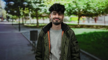 Dışarıdaki parkta gülümseyen genç adam, arka planda ağaçlar ve patika olan sıradan bir ceket giyiyor, rahat ve doğal bir atmosfer izlenimi veriyor..