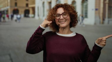 Elle işaret veren bir kadın, canlı bir şehir caddesinde duruyor, gülümsüyor ve gözlük takıyor..